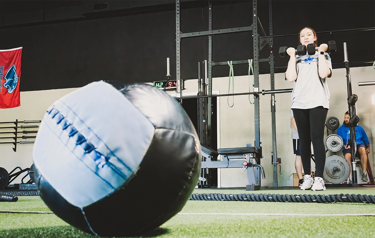 Teen athlete lifting dumbbells during training at AMP with a focus on a medicine ball during a teen athletic training program session.