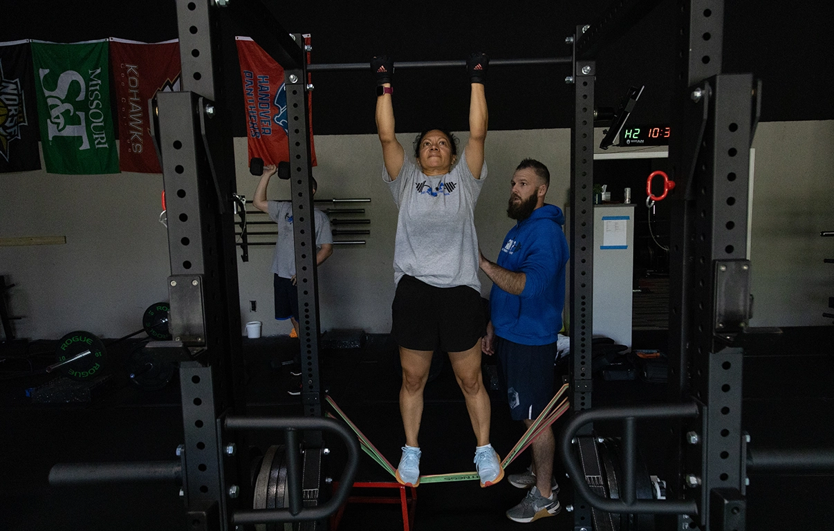 Woman doing a chin-up with Alex Makin spotting her at Alex Makin Performance for adult fitness training.