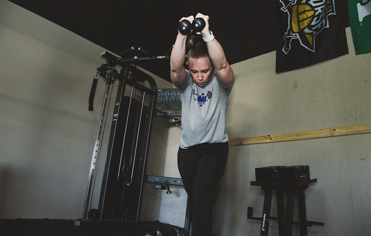 Woman lifting hand weights overhead during strength training at Alex Makin Performance's adult fitness training class.