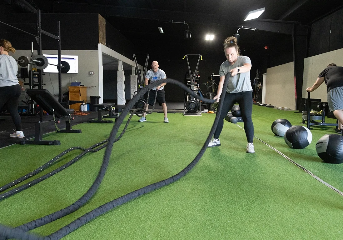 Two adults performing battle rope exercises while another pushes a weighted sled during a strength and conditioning workout with adult fitness training at Alex Makin Peformance.